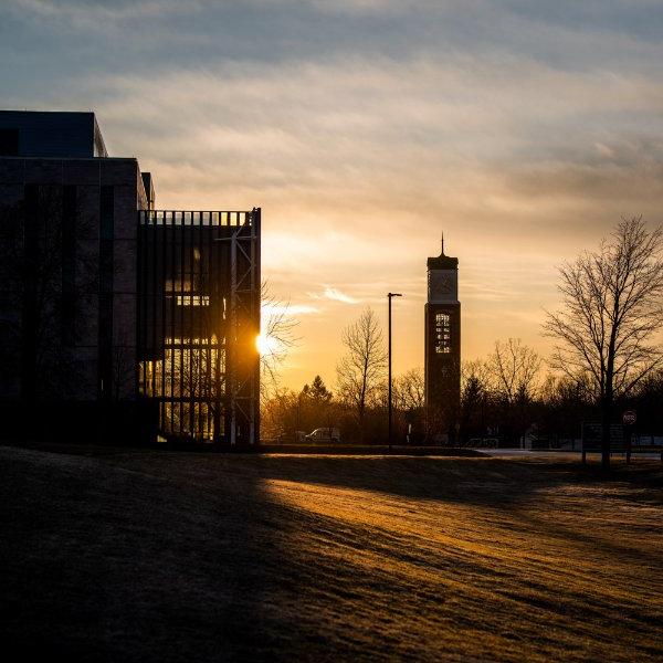 Sun rising partly behind a building, light streams across the grass and glows through the glass of the building on the right. A carillon tower is silhouetted to the left of the frame as well.