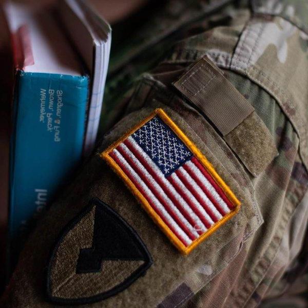 The arm of a person wearing military fatigues with a patch of the American flag holds books.